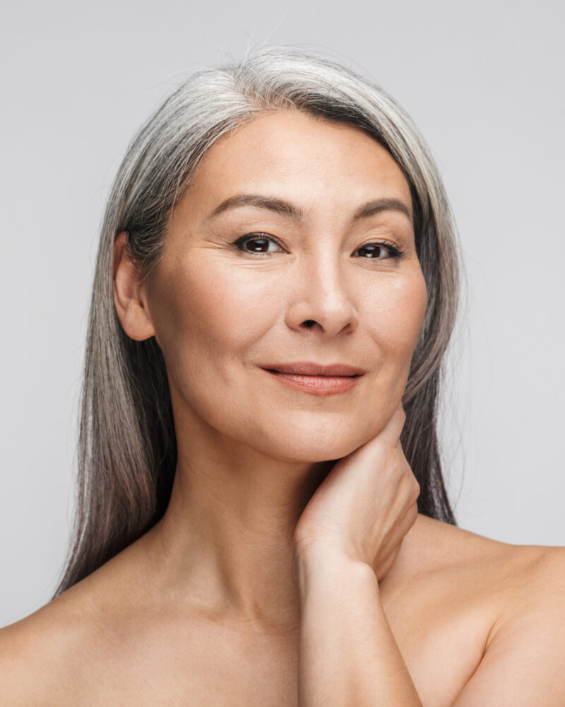 An asian woman with gray hair and glowing skin resting her hand on her neck.